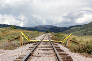 Ein Bahngleis in einer ländlichen Gegend endet abrupt mit einer kaputten Brücke, die mit gelbem Absperrband bedeckt ist. Dies schafft eine Szene, die in Fotos zum Träumen gehört. Die Gleise sind von üppigem Grün und sanften Hügeln unter einem bewölkten Himmel umgeben.