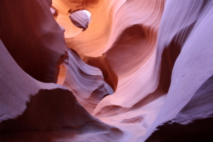 Ein schmaler Sandsteincanyon mit glatten, wellenartigen Wänden in Orange-, Rosa- und Lilatönen. Durch eine Öffnung darüber fällt Licht ein, das die Kurven und Strukturen der Felsformationen hervorhebt – wahrlich ein Anblick, der „Fotos zum Träumen“ hervorbringt.