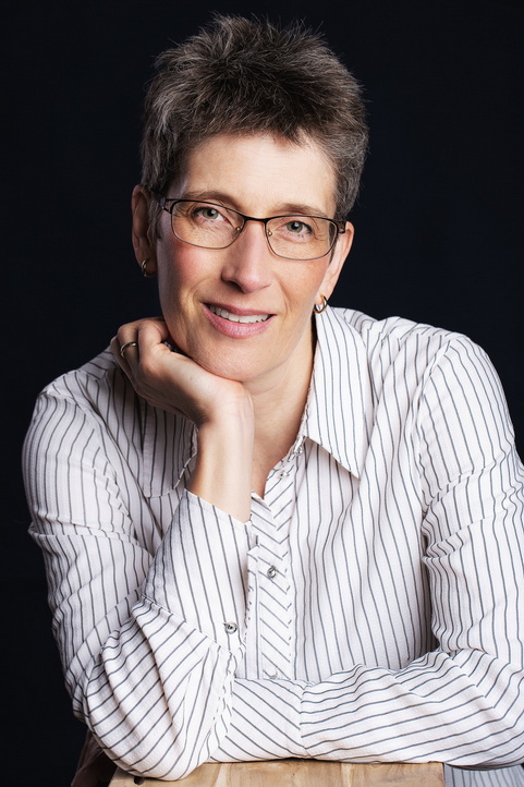 Porträt einer lächelnden Frau mit kurzen, graubraunen Haaren und Brille. Sie trägt ein weißes Hemd mit feinen, dunklen Streifen und stützt ihr Kinn auf eine Hand. Der Hintergrund ist dunkel.