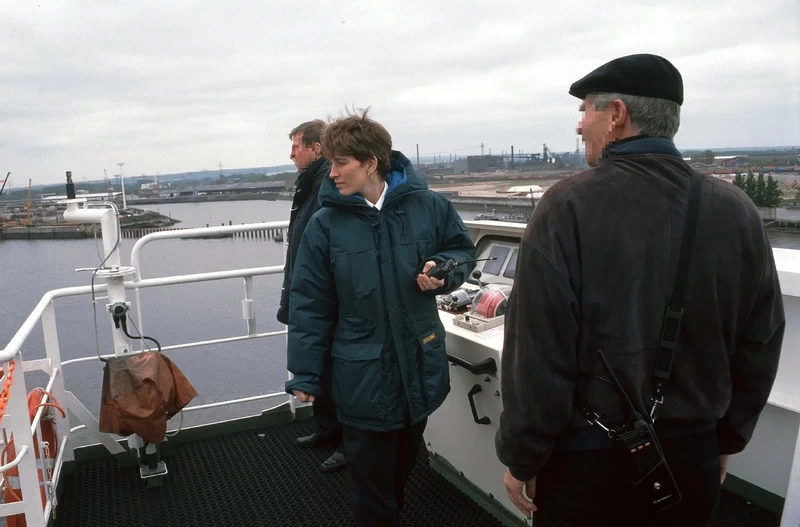 Drei Personen auf der Brücke eines Schiffes bei der Einfahrt in den Hafen von Southampton. Sabine und der Lotse schauen achteraus, Sabine hält ein Walkie-Talkie.