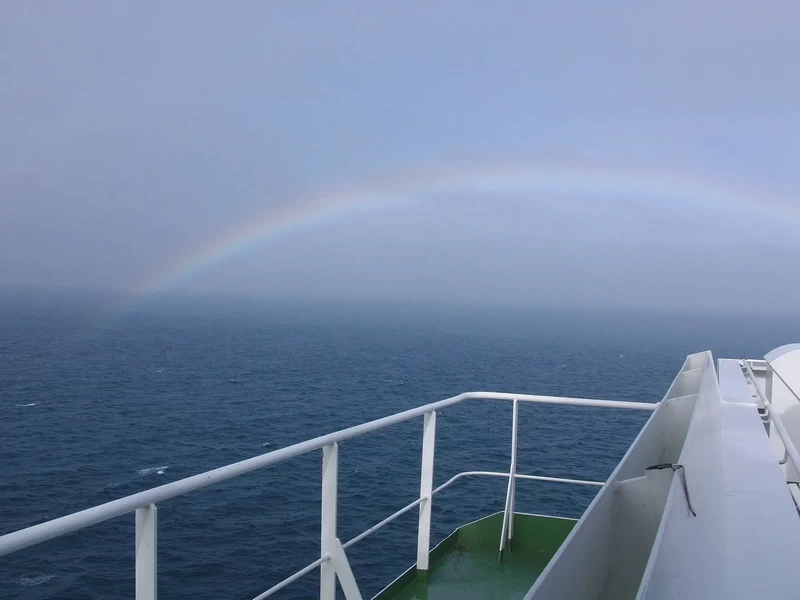 Regenbogen über dem Meer, fotografiert von der Reling eines Schiffes bei bewölktem Himmel.