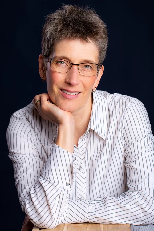 Helles Porträt von Sabine Schroeder, Autorin, mit Kurzhaarschnitt und Brille vor dunklem Hintergrund