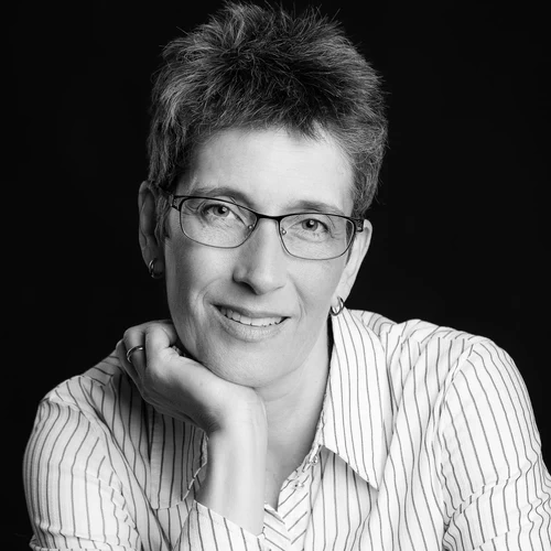 Schwarz-weiß-Porträt von Sabine Schroeder, Autorin, mit Kurzhaarschnitt und Brille vor dunklem Hintergrund (Nahaufnahme)