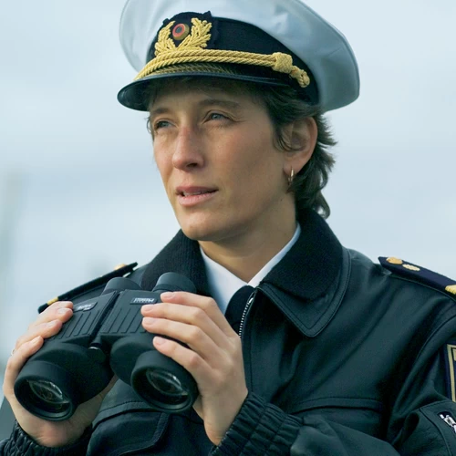Ship's officer in dark uniform with cap, gazing attentively into the distance. She holds binoculars at the ready.